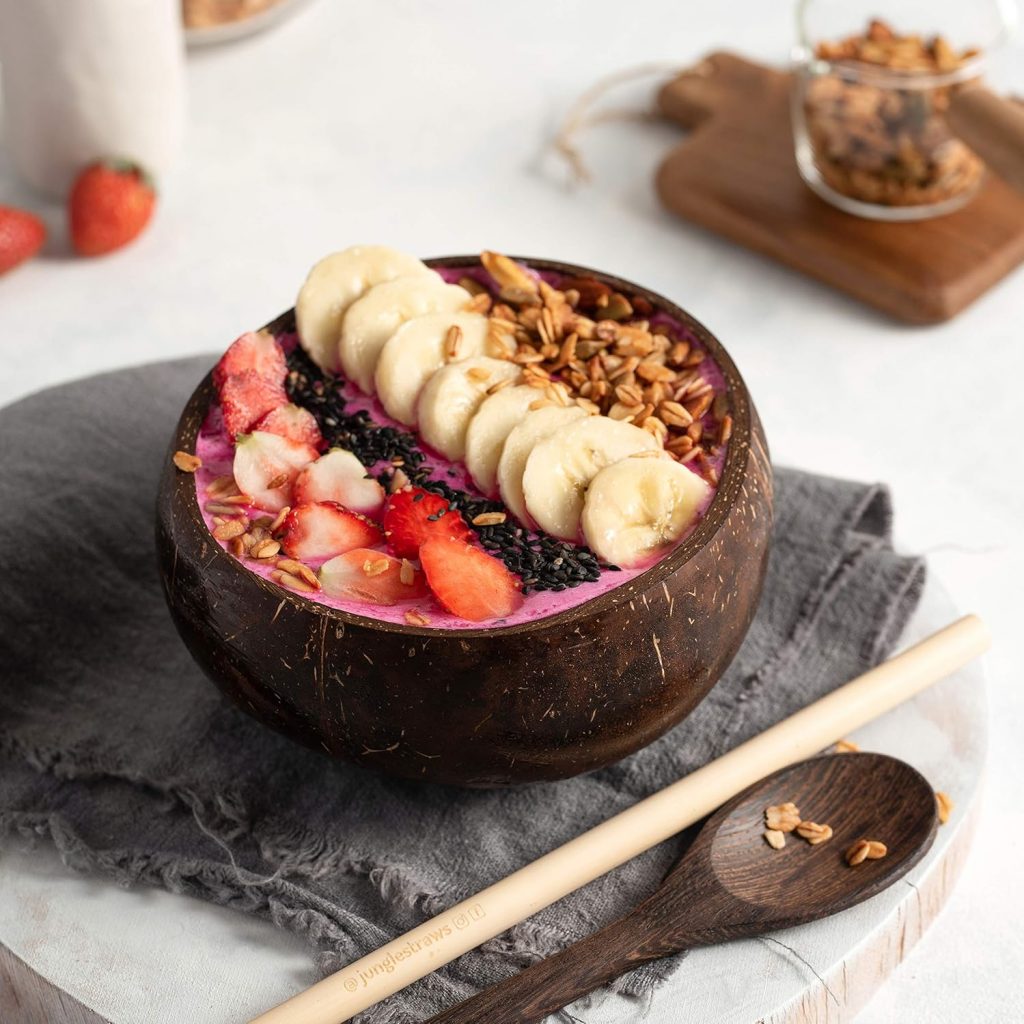 Kokosschale gefüllt mit Smoothie Bowl aus Bananen, Erdbeeren und Granola, dekorativ angerichtet auf grauer Stoffserviette mit Holzlöffel und Bambusstrohhalm – nachhaltige, natürliche Tischdeko