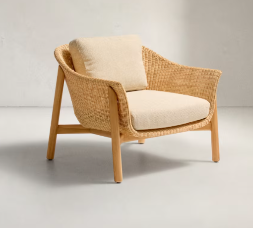 Moderner Sessel aus massiver Teakholzstruktur mit Sitzfläche und Rückenlehne aus hellem, geflochtenem Rattan und naturfarbenem Kissen – auf hellem Boden vor einer neutralen Wand platziert